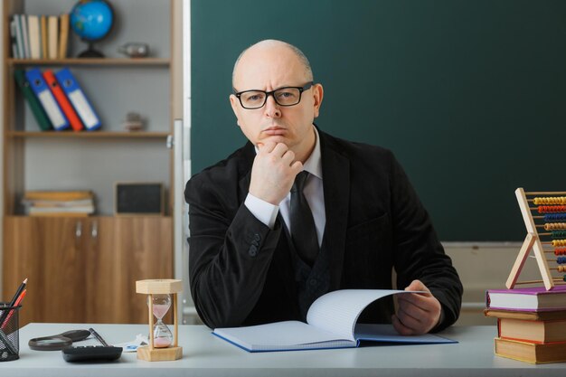 Profesor hombre con gafas revisando el registro de clase mirando a la cámara desconcertado con expresión pensativa pensando sentado en el escritorio de la escuela frente a la pizarra en el aula