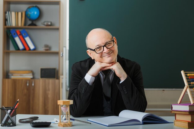 Profesor hombre con gafas revisando el registro de clase mirando a la cámara feliz y complacido sonriendo alegremente sentado en el escritorio de la escuela frente a la pizarra en el aula