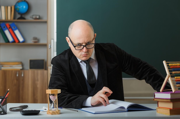 Profesor hombre con gafas revisando el registro de clase mirando a la cámara frunciendo el ceño disgustado sentado en el escritorio de la escuela frente a la pizarra en el aula