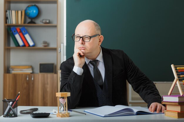 Profesor hombre con gafas revisando el registro de clase mirando a un lado con expresión pensativa pensando sentado en el escritorio de la escuela frente a la pizarra en el aula