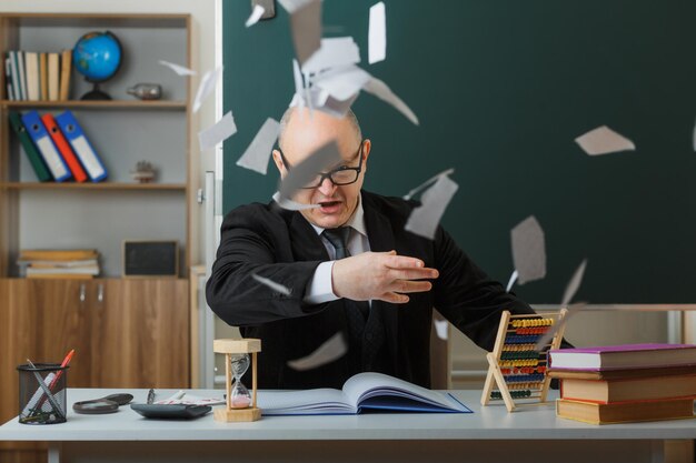 Profesor hombre con gafas sentado en el escritorio de la escuela frente a la pizarra en el aula arrojando trozos de papel con expresión agresiva enojada