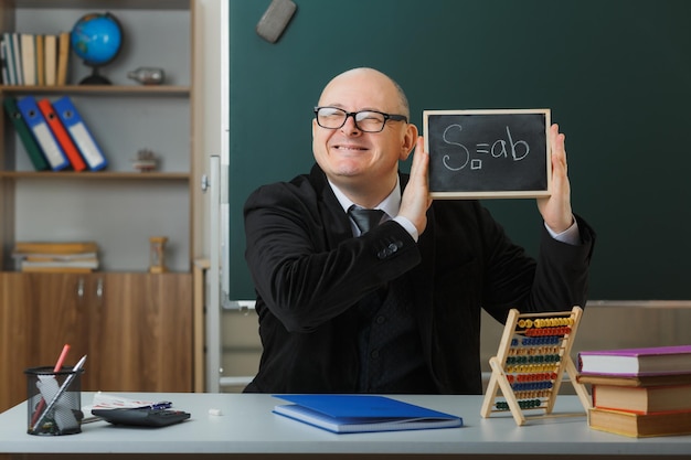Foto gratuita profesor hombre con gafas sentado en el escritorio de la escuela frente a la pizarra en el aula mostrando pizarra explicando la lección feliz y complacido sonriendo alegremente