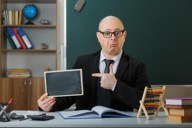 Profesor hombre con gafas sentado en el escritorio de la escuela frente a la pizarra en el aula mostrando pizarra explicando la lección señalando con el dedo índice intrigado