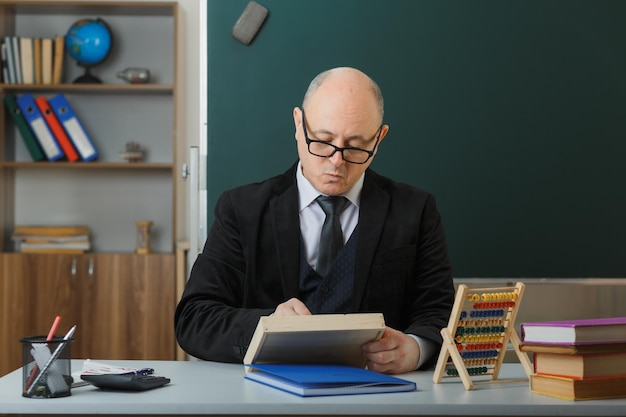 Profesor hombre con gafas sentado en el escritorio de la escuela frente a la pizarra en el aula preparándose para la lección con cara seria