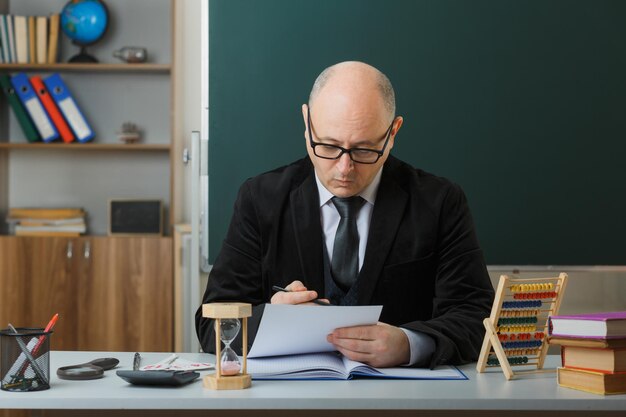 Profesor hombre con gafas sentado en el escritorio de la escuela frente a la pizarra en el aula revisando la tarea de los estudiantes con cara seria concentrada