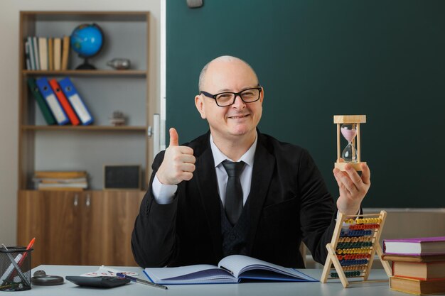 Profesor hombre con gafas sentado en el escritorio de la escuela frente a la pizarra en el aula sosteniendo un reloj de arena explicando la lección mostrando el pulgar hacia arriba feliz y complacido