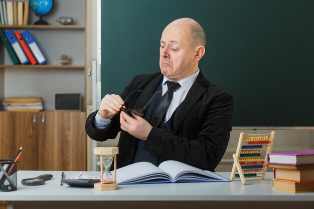 Profesor hombre con gafas sentado en el escritorio de la escuela con registro de clase frente a la pizarra en el aula explicando la lección con confianza