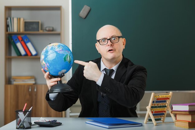 Profesor hombre con gafas sentado con globo en el escritorio de la escuela frente a la pizarra en el aula explicando la lección apuntando con el dedo índice al globo mirando a la cámara