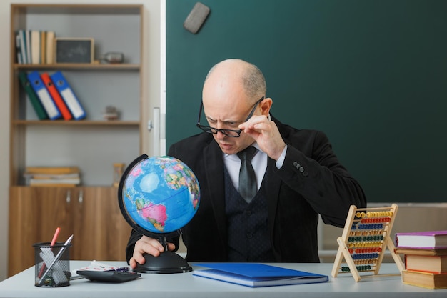 Profesor hombre con gafas sentado con globo en el escritorio de la escuela frente a la pizarra en el aula explicando la lección confundido