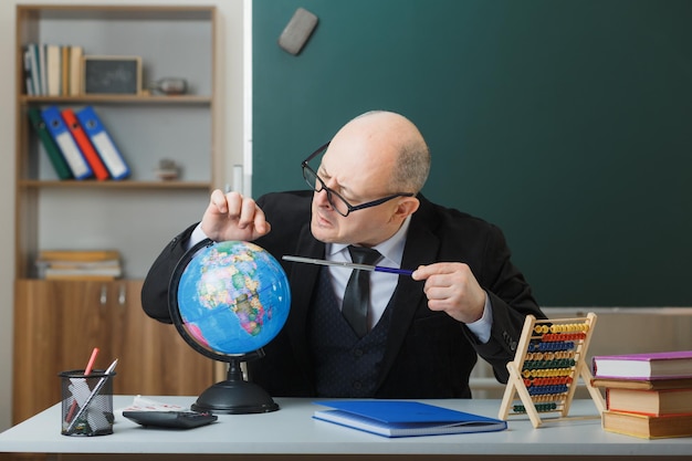 Profesor hombre con gafas sentado con globo en el escritorio de la escuela frente a la pizarra en el aula explicando la lección sosteniendo el puntero mirando intrigado