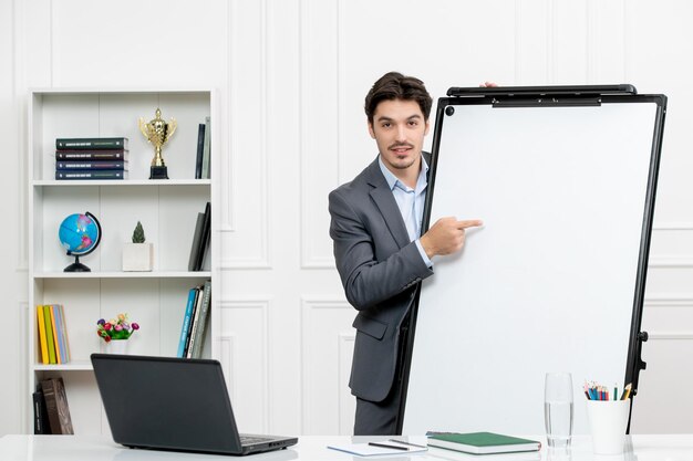 Profesor instructor inteligente en traje gris en el aula con computadora y pizarra apuntando a la pizarra