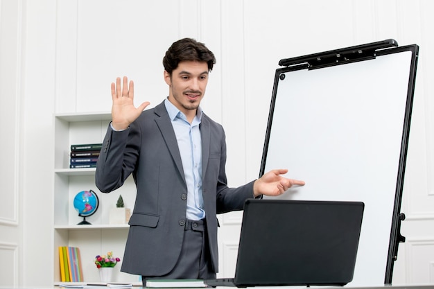 Profesor instructor inteligente en traje gris en el aula con computadora y pizarra diciendo adiós