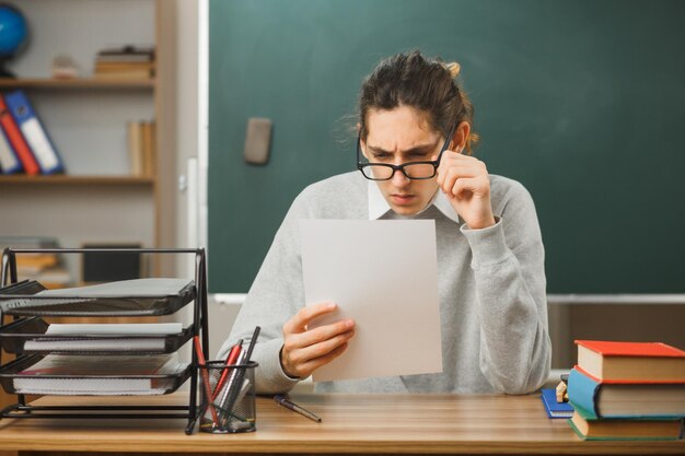 un profesor joven y estricto con gafas sosteniendo y mirando papel sentado en el escritorio con herramientas escolares en el aula