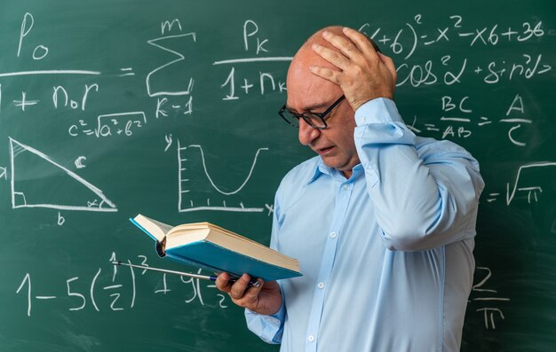 Profesor de mediana edad asustado con gafas de pie delante de la pizarra leyendo el libro poniendo la mano en la cabeza