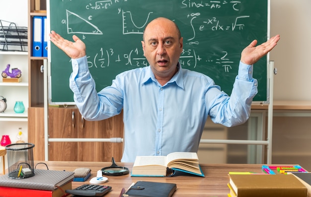 Profesor de mediana edad confundido se sienta a la mesa con herramientas escolares extendiendo las manos en el aula