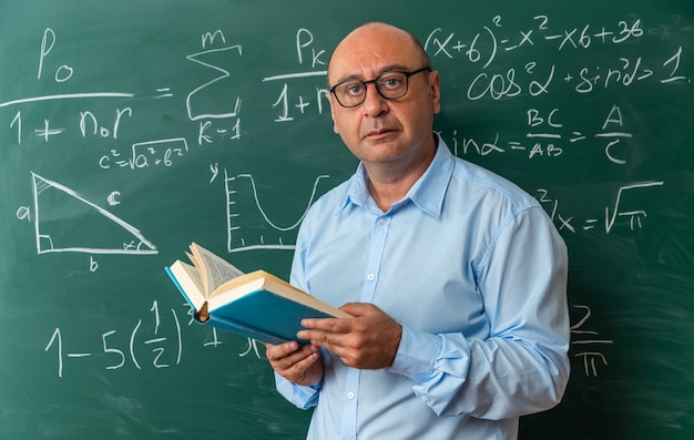 Profesor de mediana edad mirando cámara confidente con gafas de pie delante de la pizarra sosteniendo el libro