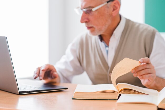 Profesor senior trabajando con laptop mientras sostiene la página del libro