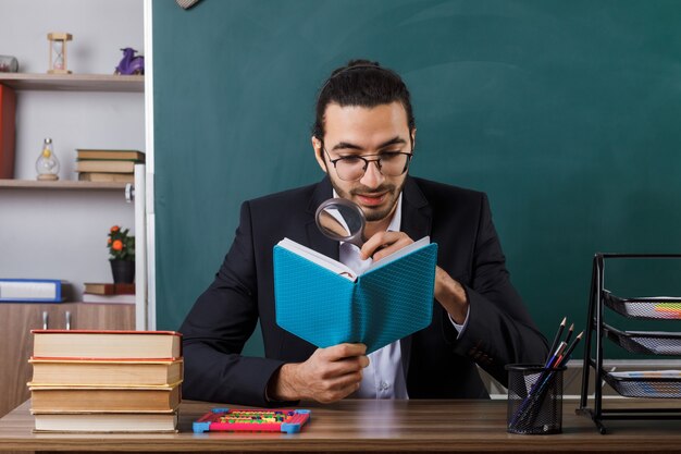 Profesor de sexo masculino complacido con gafas sosteniendo y leyendo un libro con lupa sentado a la mesa con herramientas escolares en el aula