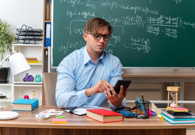 Profesor de sexo masculino joven con gafas escribiendo mensajes con smartphone mirando confiado sentado en el escritorio de la escuela con libros y notas frente a la pizarra en el aula