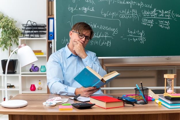 Profesor de sexo masculino joven con gafas leyendo con libro de cara seria preparando la lección sentado en el escritorio de la escuela con libros y notas frente a la pizarra en el aula