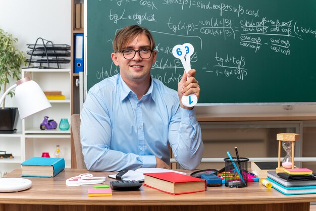 Profesor de sexo masculino joven con gafas mostrando placas de matrícula explicando la lección sonriendo sentado en el escritorio de la escuela con libros y notas frente a la pizarra en el aula