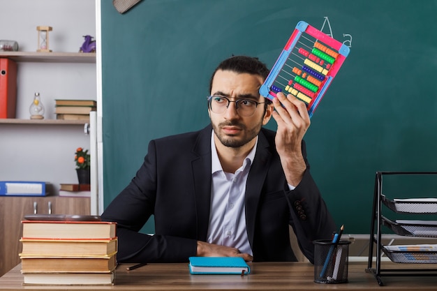 Foto gratuita profesor de sexo masculino de lado mirando disgustado con gafas sosteniendo abacus sentado a la mesa con herramientas escolares en el aula