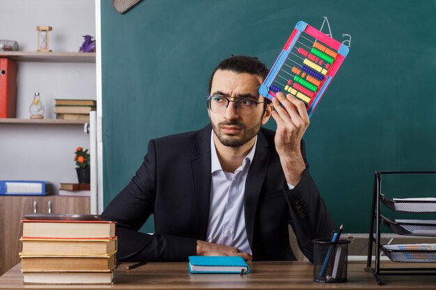 Profesor de sexo masculino de lado mirando disgustado con gafas sosteniendo abacus sentado a la mesa con herramientas escolares en el aula