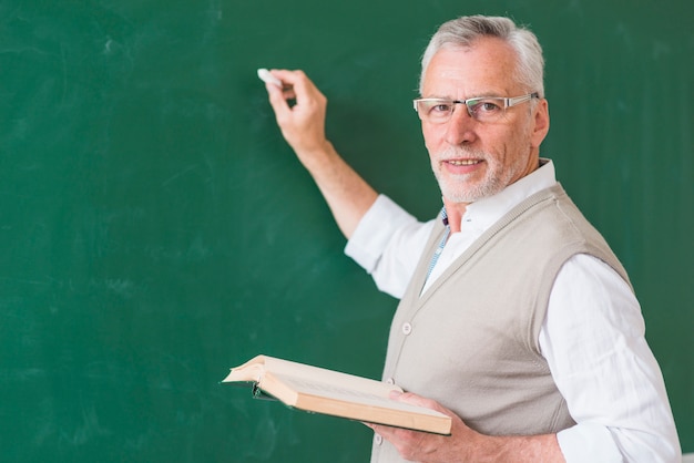 Profesor de sexo masculino mayor que sostiene el libro y que escribe en la pizarra