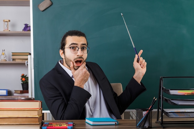 Profesor de sexo masculino de mentón agarrado asustado con puntos de gafas con puntero en la pizarra sentado a la mesa con herramientas escolares en el aula
