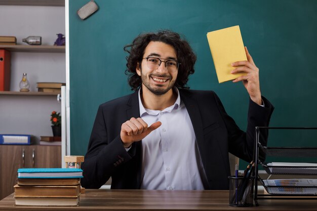 Profesor sonriente con gafas sosteniendo y apunta al libro sentado a la mesa con herramientas escolares en el aula