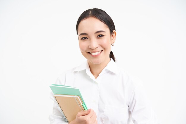 Profesora asiática con cuadernos sonriendo y luciendo segura aislada de fondo blanco