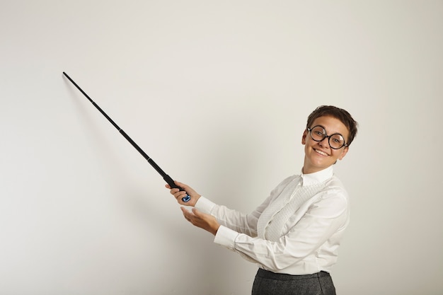 Profesora caucásica vestida de forma conservadora en feos vasos redondos sosteniendo un puntero a una pizarra vacía y sonriendo desagradablemente