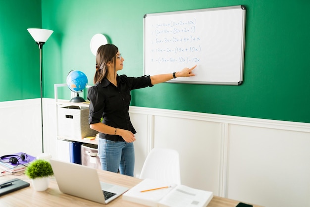 Profesora hispana dando una lección de matemáticas y escribiendo ecuaciones en la pizarra durante una clase virtual