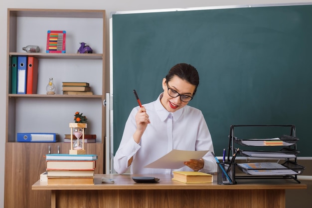 Profesora joven con anteojos revisando la tarea de los estudiantes sonriendo feliz y positiva sentada en el escritorio de la escuela frente a la pizarra en el aula