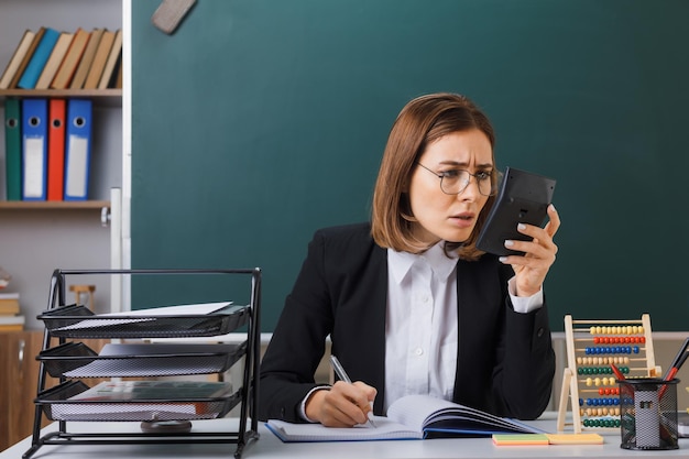 Profesora joven con anteojos sentada en el escritorio de la escuela frente a la pizarra en el aula usando una calculadora preparándose para la escritura de la lección en el registro de clase con cara tensa