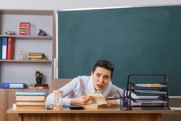 Profesora joven con anteojos sentada en el escritorio de la escuela con un libro frente a la pizarra en el aula que parece confundida mientras explica la lección