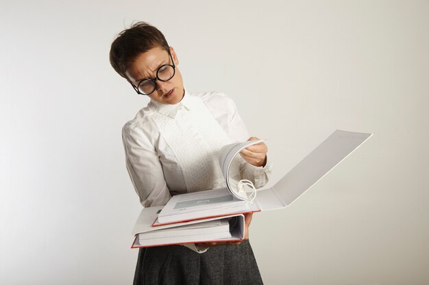 Profesora joven cansada y con el ceño fruncido en traje conservador y gafas redondas negras pasando páginas en una carpeta gruesa en blanco