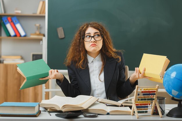 profesora joven confundida con gafas sosteniendo libros sentados en el escritorio con herramientas escolares en el aula