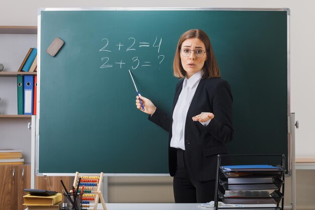 Profesora joven con gafas de pie cerca de la pizarra en el aula explicando la lección apuntando a la pizarra con el puntero mirando confundido