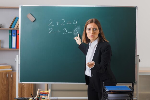 Profesora joven con gafas de pie cerca de la pizarra en el aula explicando la lección apuntando a la pizarra con un puntero que parece decepcionado