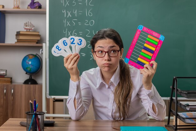 Profesora joven con gafas sentado en el escritorio de la escuela con facturas y matrículas con aspecto cansado y aburrido soplar las mejillas explicando la lección frente a la pizarra en el aula