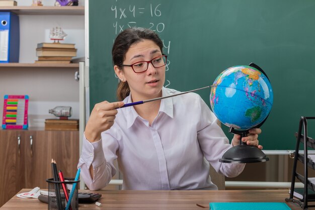 Profesora joven con gafas sosteniendo globo y puntero explicando la lección mirando confiado sentado en el escritorio de la escuela frente a la pizarra en el aula