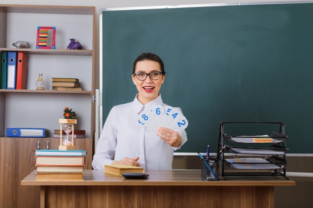 Profesora joven con gafas sosteniendo matrículas explicando la lección sonriendo confiada sentada en el escritorio de la escuela frente a la pizarra en el aula