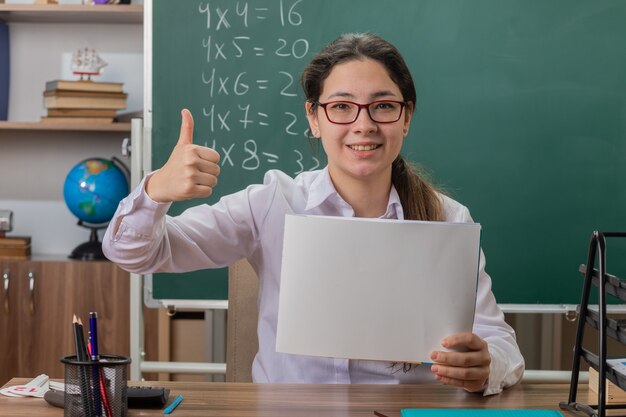 Profesora joven con gafas sosteniendo páginas en blanco mirando al frente sonriendo alegremente mostrando los pulgares para arriba sentado en el escritorio de la escuela frente a la pizarra en el aula