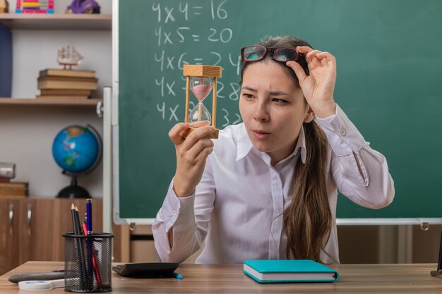 Profesora joven con reloj de arena mirándolo de cerca preparándose para la lección sentado en el escritorio de la escuela frente a la pizarra en el aula