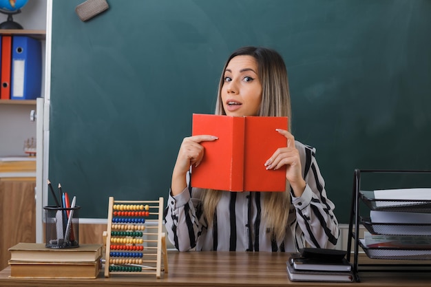 profesora joven sentada en el escritorio de la escuela frente a la pizarra en el aula sosteniendo un libro mirando a la cámara con una expresión astuta