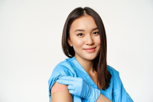 Programa de vacunación y concepto de covid-19. Retrato de trabajador de la salud asiático en bata médica, mostrando el brazo vacunado y sonriendo, de pie sobre fondo blanco.