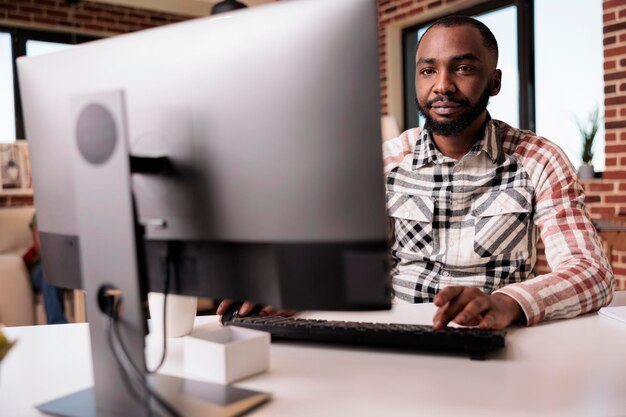 Programador afroamericano confiado mirando la pantalla de la computadora y escribiendo mientras trabaja a distancia desde la sala de estar de su casa. Enfoque selectivo en autónomos que usan pc para trabajo remoto sentado en el escritorio.