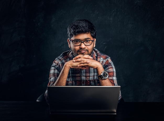 El programador indio se sienta en un escritorio con una computadora portátil y mira la cámara con una mirada pensativa. Foto de estudio contra una pared de textura oscura.