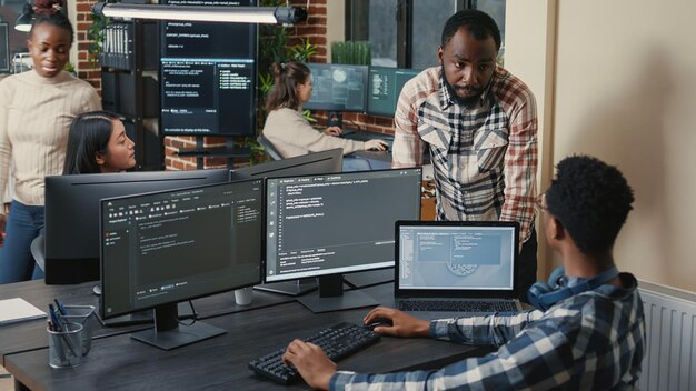 Programador sentado en el escritorio con varias pantallas ejecutando código hablando con un colega sobre el algoritmo de inteligencia artificial. Desarrolladores de software que realizan un proyecto innovador de inteligencia artificial.